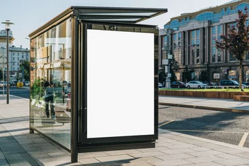 Fotobehang Verticaal leeg wit reclamebord bij bushalte op stadsstraat. Op de achtergrond gebouwen en weg. Bespotten. Affiche op straat naast rijbaan. Zonnige zomerdag. © foxyburrow