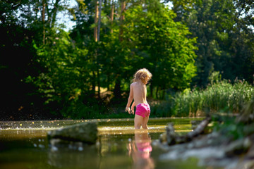 Adorable curly toddler girl wearing in pink shorts, playing at a river shore splashing water on sunny summer day