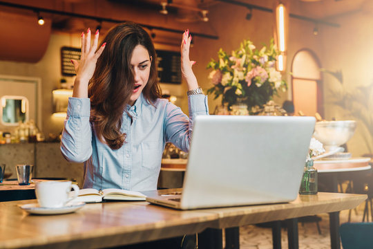 Young businesswoman is sitting in cafe at table in front of laptop and opening her mouth and raising her hands in surprise looks at screen. Girl received an email and is happy with good news.