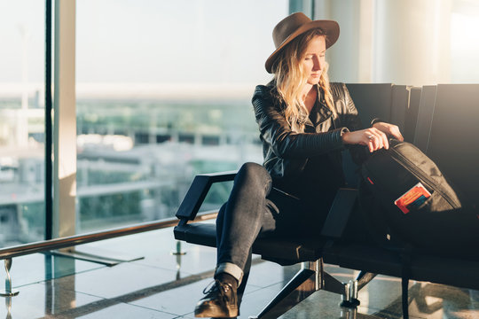 Young tourist woman in hat sits at airport near window and is looking for something in her backpack. Hipster girl is flying on vacation, traveling. Lifestyle, leisure, relaxing. Blurred background.