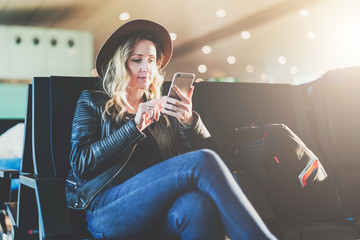Young woman tourist in hat, with backpack sits at airport and uses smartphone. Hipster girl is waiting for plane landing, checks email, chatting, blogging, browsing internet.