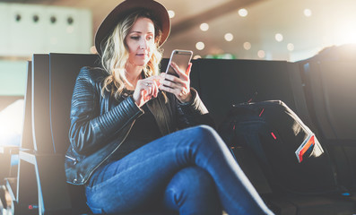 Young woman tourist in hat, with backpack sits at airport and uses smartphone. Hipster girl is waiting for plane landing, checks email, chatting, blogging, browsing internet.