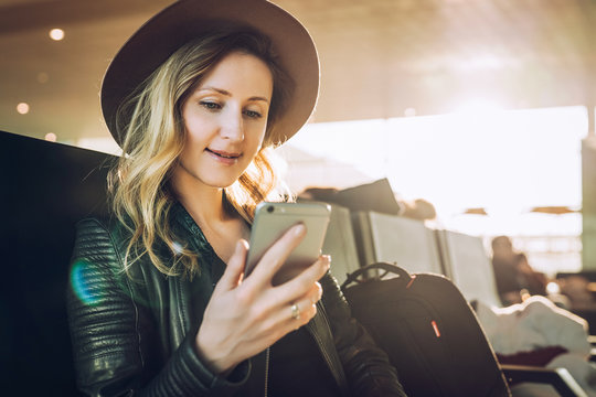 Young woman tourist in hat, with backpack sits at airport and uses smartphone. Hipster girl is waiting for plane landing, checks email, chatting, blogging, browsing internet.