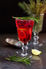 a glass with a cocktail of red color with lime, rosemary and ice cubes on a background of rosemary on a dark background