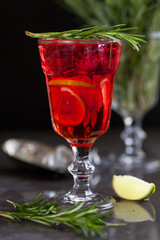 a glass with a cocktail of red color with lime, rosemary and ice cubes on a background of rosemary on a dark background