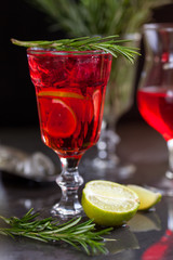 a glass with a cocktail of red color with lime, rosemary and ice cubes on a background of rosemary on a dark background
