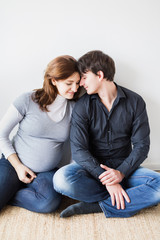 pregnancy, parenthood, fashion concept. on the floor by the white wall there is a man and a woman dressed in casual style, she is dressed in grey t shirt and he is in black shirt