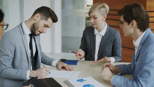 Young businessman in suit signing contract and shake hands with his colleague while corporate lawyer explaining details of partnership sitting in modern office