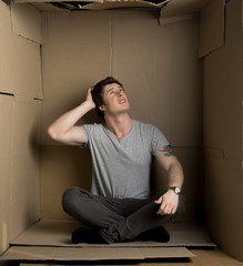 Problem of space concept. Full length of young man is expressing concern while looking at paper ceiling of his carton box and touching his head. He is feeling discomfort while being in small housing