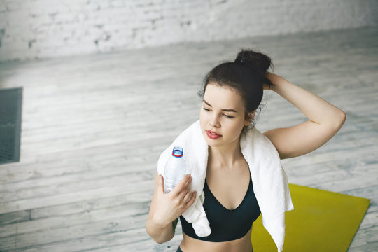 Close up shot of beautiful tired young female with white towel around her neck holding plastic bottle of water, refreshing herself after exhaustible cardio workout in gym hall with copy space floor