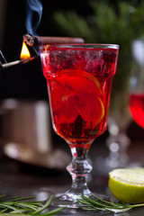 a glass with a cocktail of red color with lime, red berries, rosemary, burning cinnamon and ice cubes on a background of rosemary on a dark background