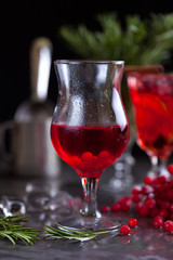 a glass with a cocktail of red color with lime, red berries, rosemary and ice cubes on a background of rosemary on a dark background