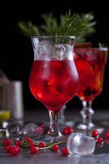 a glass with a cocktail of red color with lime, red berries, rosemary and ice cubes on a background of rosemary on a dark background