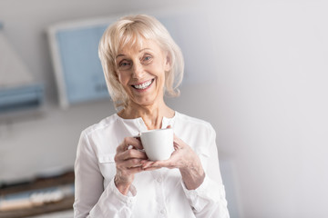 Morning coffee. Jolly merry mature woman looking at camera while carrying cup and smiling