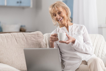 Funny comedy. Glad positive mature woman staring at screen while drinking coffee and laughing