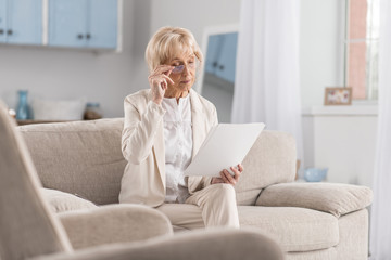 Woman in business. Appealing earnest mature businesswoman sitting on couch while working with papers and touching glasses