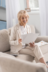 Precise look. Positive optimistic mature businesswoman sitting on couch while carrying papers and smiling
