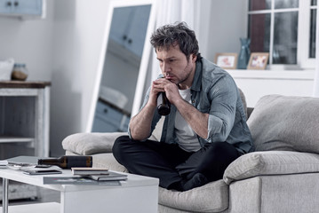 Terrible addiction. Unhappy depressed man sitting on a sofa with his legs crossed and holding a bottle while sadly thinking about his terrible alcohol addiction