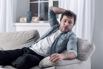 Fototapeta premium Lazy man. Calm unshaved unemployed lazy man sitting comfortably on a sofa and relaxing instead of finding a new well paid job