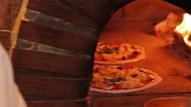 baking traditional italian pizza in a wood fired stone oven with a lot of toppings. pizzaiolo is using a pizza peel.