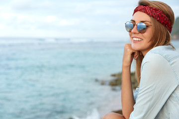 Glad young cute female hiker wears sunglasses and headband, sits on cliff, looks at beautiful view of peaceful ocean or sea with copy space for your text or advertisment. Seascape and nature.