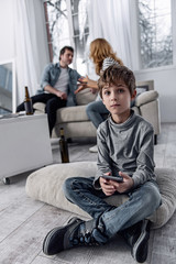 Unpleasant atmosphere. Upset little boy feeling unhappy and resting on the pillow with a smartphone in his hands while his parents sitting on the sofa behind his back and discussing their divorce