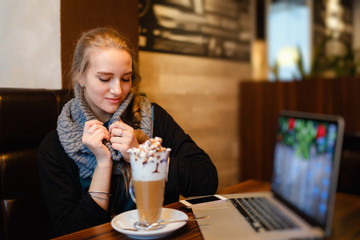 A young girl drinks cappuccino with marshmallow working at the laptop sitting in a cozy warm cafe. Girl warm warm coffee after street.