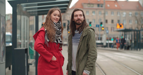 Smiling couple looking away while waiting at tram stop in a city.