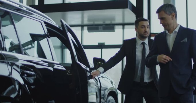 Elegant salesman inviting potential buyer to get in the car in showroom. Slow motion. Shot on Red cinema camera.