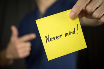 Man holds yellow sticker inscription Never mind!. Thumb up Blue T-shirt. close-up, selective focus
