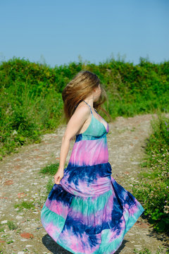 white caucasian blonde girl with colorful long dress dances on a dirt road in field