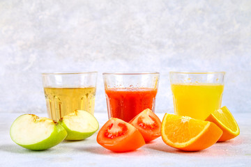 Glasses with fresh orange, apple, tomato juice on a gray concrete table. Lobules Fruits and vegetables around.