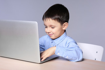 Cute little boy using laptop while doing homework against grey background