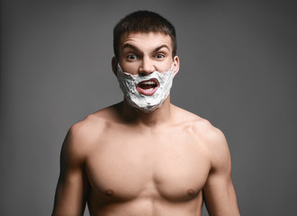 Emotional young man with shaving foam on his face against grey background