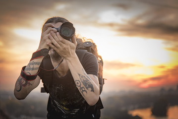 Young photographer with long hair and alternative style taking photographs with his dslr camera, capturing landscape and sunset in a park