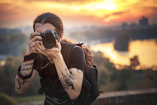 Young photographer with long hair and alternative style taking photographs with his dslr camera, capturing landscape and sunset in a park