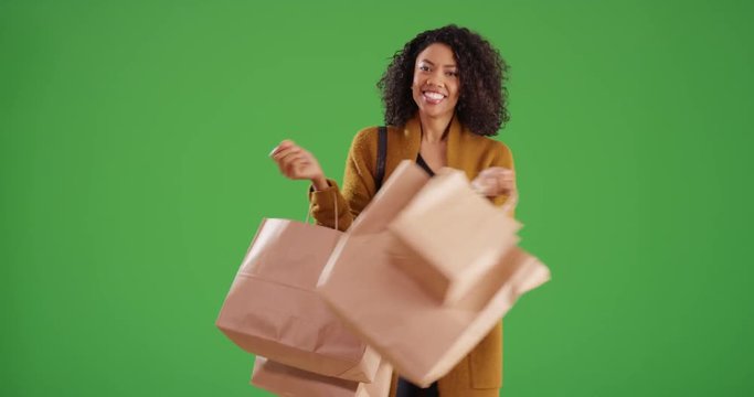 Happy stylish African American woman spinning while holding shopping bags on greenscreen. Fashionable black millennial shopper dances in yellow coat on green screen for keying or compositing. 4k