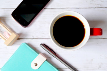 Selective focus and top view of black coffee in red mug with notebook,pen,hourglass and smartphone on white wooden background.