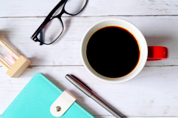 Selective focus and top view of black coffee in red mug with notebook,pen,hourglass and sunglasses on white wooden background.