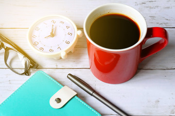 Selective focus and top view of black coffee in red mug with notebook,pen,clock and hourglass on white wooden background.