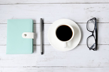 Top view and selective focus of a cup of black coffee with sunglasses, notebook and pen on white wooden background.