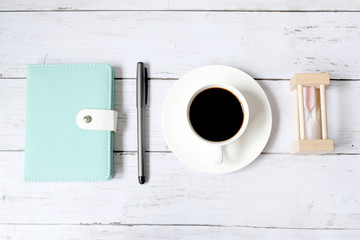 Top view and selective focus of a cup of black coffee with hourglass, notebook and pen on white wooden background.