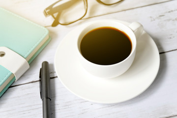 Top view and selective focus of a cup of black coffee with sunglasses, notebook and pen on white wooden background.