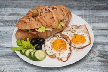 Croissant Sandwich with Fried Eggs and Fresh Vegetables on White Plate over Gray rustic Wooden Background.