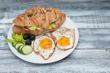 A plate of Fried Eggs with Sandwich Croissant, Cucumbers, Black Olives on Rustic Gray Background. Top view. Morning Food. copy space for Text.