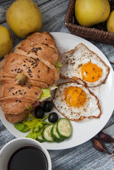Croissant Sandwich with Fried Eggs, cucumbers and olives, Fruits and Fresh Vegetables and Cup of coffee on White Plate over Gray rustic Wooden Background. Vertical Image.