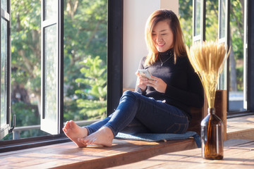 Asian woman relaxing and using her smartphone with dry flower on wooden table near the window in the room