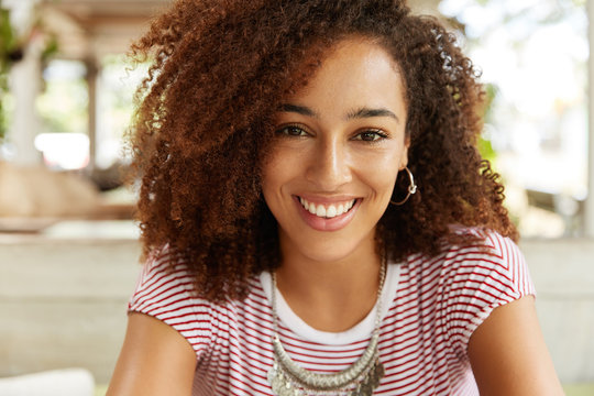 Close up shot of attractive dark skinned female with broad smile, dressed in striped t shirt, rests at home, happy to have day off, has happy expression. Smiling African American woman poses indoor