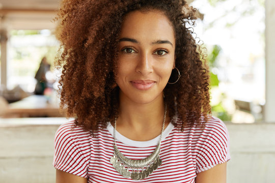 Photo of pretty African American female in striped t shirt, has positive expression, listens interlocutor who sits opposite her, spend dinner break at cafeteria. People, beauty and lifestyle concept