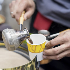 Close-up of hands of man making traditional turkish coffee in copper turk on hot sand and pours into a disposable paper cup. Coffee preparation concept